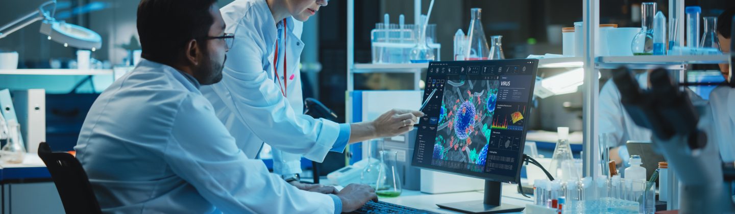 Female Research Scientist With Bioengineer Working On A Personal Computer With Screen Showing Virus Analysis Software User Interface. Scientists Developing Vaccine, Drugs And Antibiotics In Laboratory